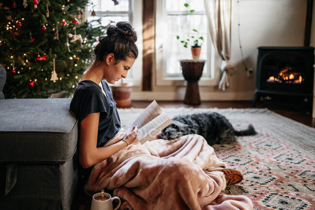 Young woman reading with dog, sitting by fireplace and Christmas tree — Stock Photo