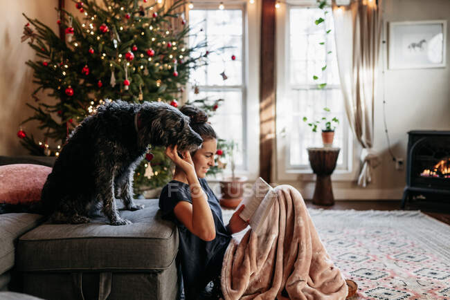Feliz adolescente leitura por lareira e árvore de Natal, acariciando seu cão — Fotografia de Stock