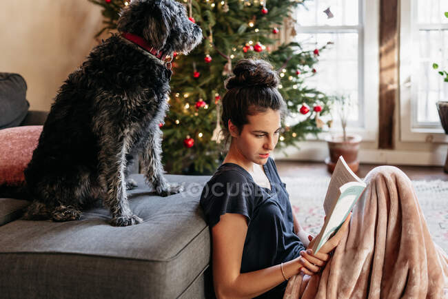 Jeune femme lisant avec son chien, sapin de Noël en arrière-plan — Photo de stock