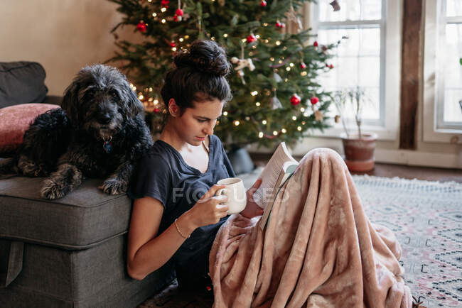 Jovem mulher lendo com seu cão, árvore de Natal no fundo — Fotografia de Stock