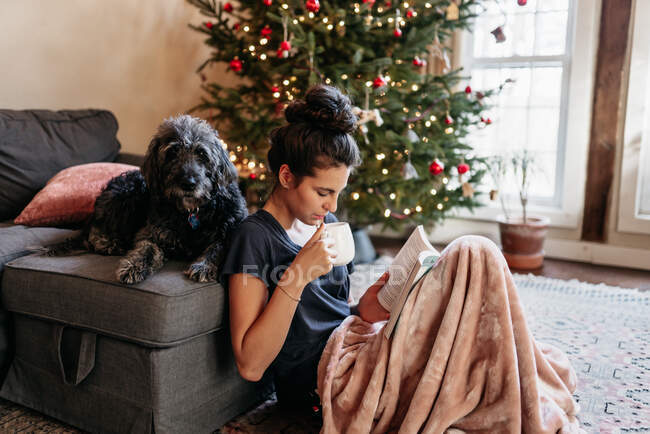 Young woman reading and drinking tea with dog bu Christmas tree — Stock Photo