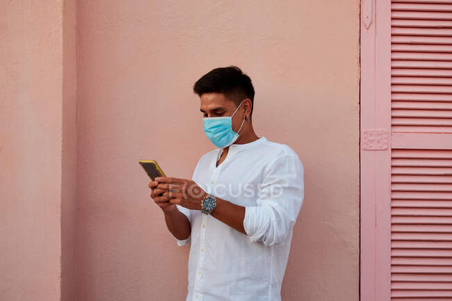 Молодий латинський чоловік з маскою дивиться свій телефон на рожевий фон — стокове фото