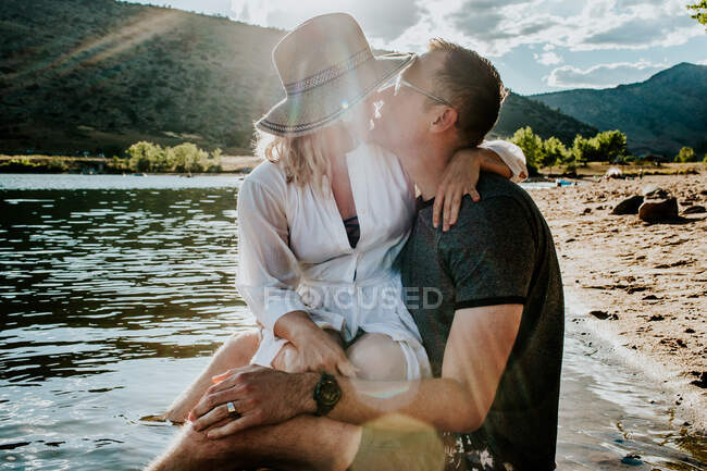 Marido y mujer sentados juntos y besándose en la orilla del lago - foto de stock