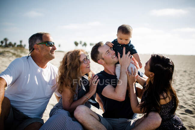 Fünfköpfige Familie sitzt am Strand und lächelt Säugling an — Stockfoto