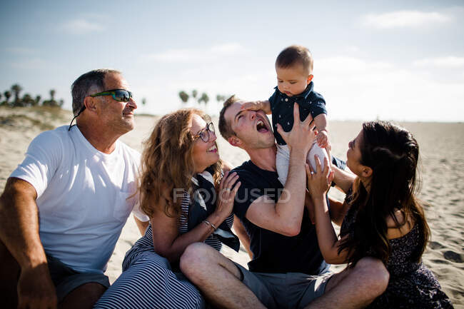 Bebé aplastando a papá en la cara, sentado en la playa con la familia sonriendo - foto de stock