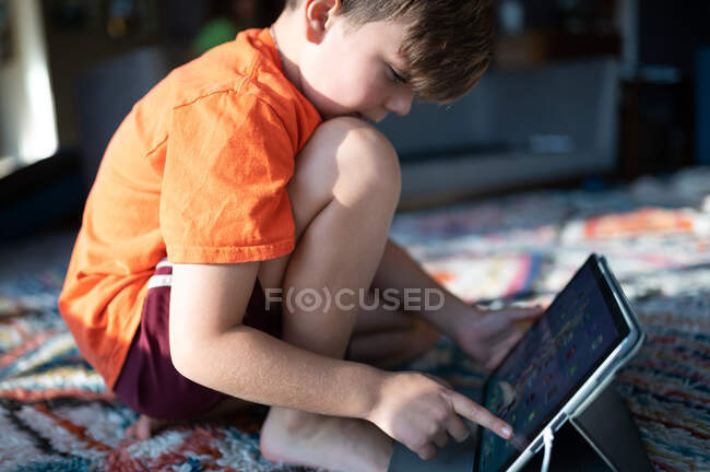 Niño arrodillado en la alfombra en casa con ipad tableta de selección de aplicaciones - foto de stock