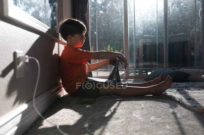 Boy sitting on the floor with ipad tablet plugged in to the wall — Stock Photo