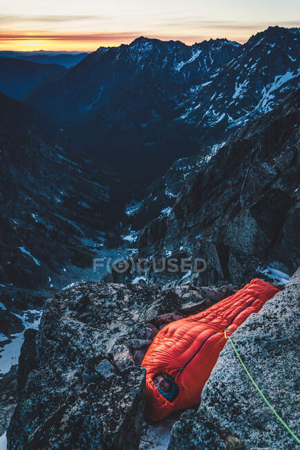 5 minutos más. hombre disfruta de la comodidad de un saco de dormir caliente mientras está atado en el ancla en un panal expuesto en el monte. Stuart, Washington. - foto de stock