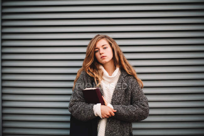 Портрет милой студентки-подростка с книгой, стоящей у серой стены — стоковое фото