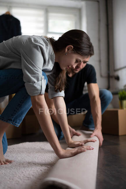Vista laterale della giovane signora in abito casual sorridente e srotolamento tappeto bianco sul pavimento con l'aiuto del fidanzato nel nuovo appartamento — Foto stock