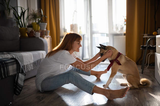 Side view of delighted barefoot female smiling and playing with dog while sitting on floor at home — Stock Photo