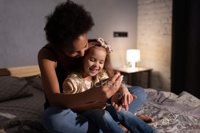 Этническая женщина и жизнерадостная девочка смешанной расы улыбаются и играют с маленькими игрушками, сидя вечером дома на кровати — стоковое фото