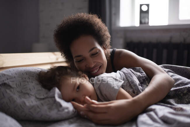 Encantada mujer étnica sonriendo y abrazando linda chica de raza mixta mientras está acostada en la cama y despertando a su hija por la mañana - foto de stock