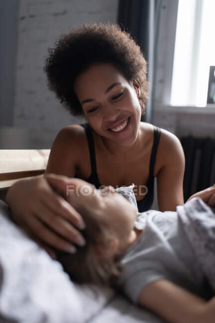 Cheerful mixed race woman smiling and stroking head of girl while waking up daughter in morning in cozy bedroom — Stock Photo