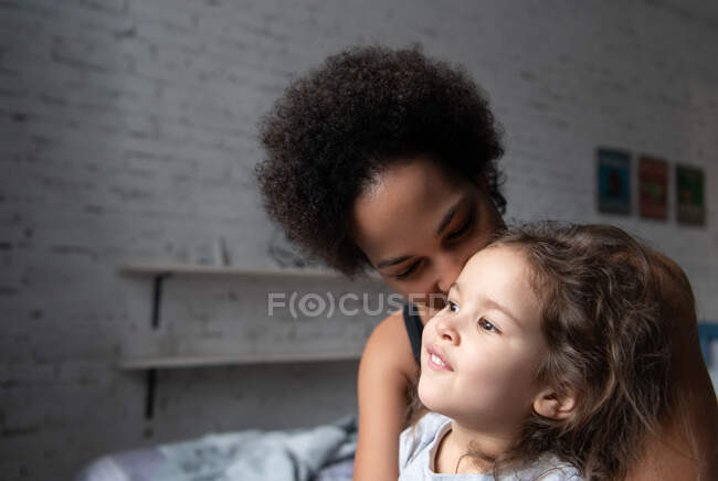 Etnia mulher abraçando e beijando bonito misto raça menina enquanto sentado na cama no aconchegante quarto em casa — Fotografia de Stock
