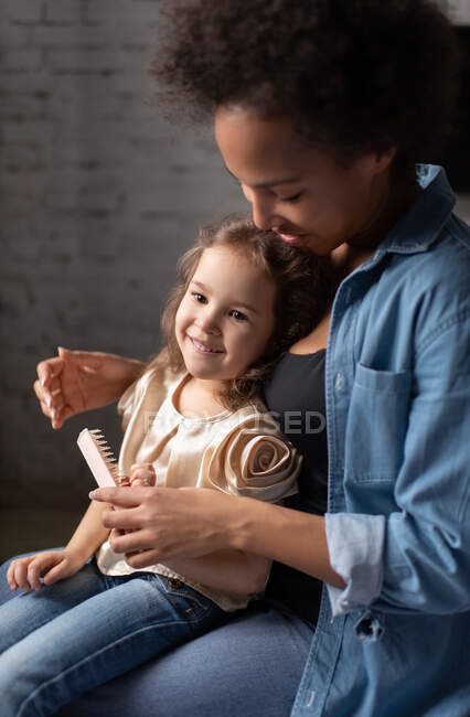 Позитивная этническая женщина с расческой учит счастливую смешанную расовую девушку расчесывать волосы, проводя время дома вместе — стоковое фото