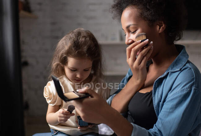 Madre étnica usando cepillo para esparcir polvo en la cara mientras enseña a la niña de raza mixta a aplicar maquillaje en casa - foto de stock