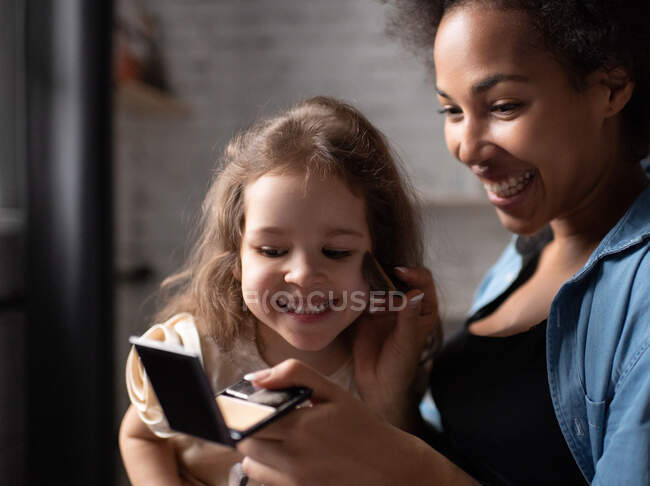 Delighted ethnic mother with compact spreading powder on face of smiling mixed race girl while teaching daughter to apply makeup at home — Stock Photo