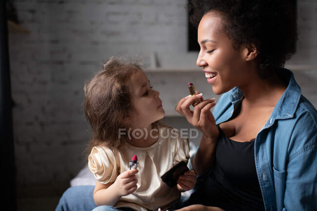 Délicieuse femme ethnique souriante et montrant fille mixte mignonne comment appliquer du rouge à lèvres pendant la leçon de maquillage à la maison — Photo de stock