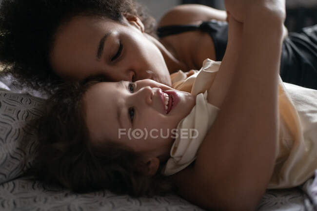 Hochwinkel der ethnischen Frau mit geschlossenen Augen küsst fröhliche gemischte Rassenmädchen in die Wange, während sie mit Tochter auf dem Bett spielt — Stockfoto