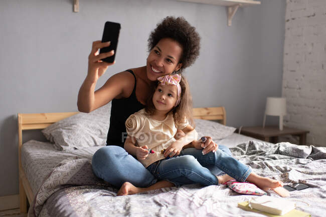 Mujer étnica feliz sonriendo y abrazando linda chica de raza mixta mientras está sentado en la cama y el uso de teléfono inteligente para hacer videollamada a la familia - foto de stock