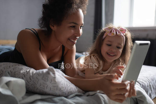 Entzückte Frau und Mädchen liegen im Bett und spielen gemeinsam am Wochenende zu Hause ein Spiel auf dem digitalen Tablet — Stockfoto