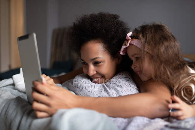Alegre mujer de raza mixta y niña sonriendo y utilizando la tableta para hacer videollamada a los familiares mientras está acostado en la cama en casa - foto de stock
