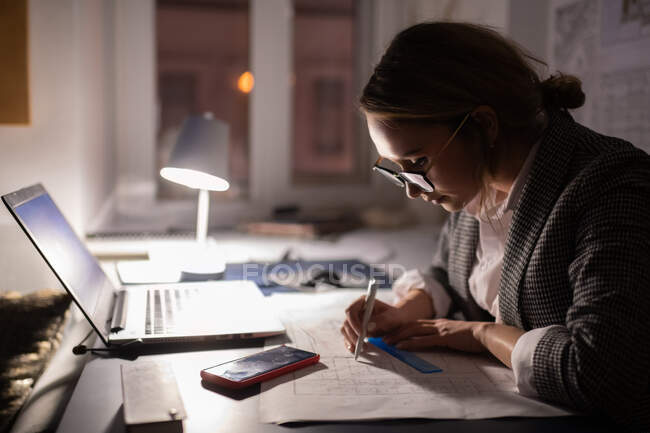 Вид сбоку на молодую деловую женщину в очках, рисующую черновой вариант, сидя за столом с ноутбуком и смартфоном во время работы на темном рабочем месте ночью — стоковое фото