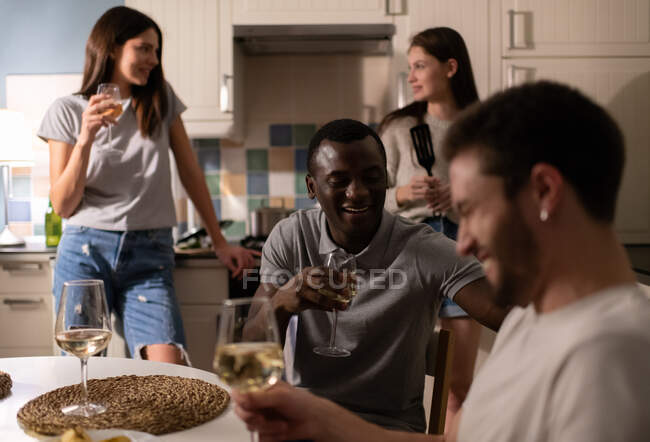 Cheerful African American guy drinking wine and speaking with laughing friend while sitting at table near cooking girlfriends — Stock Photo