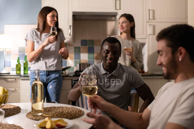 Schwarzer Mann lächelt und prostet optimistischem Mann zu, während er am Tisch neben kochenden Freundinnen sitzt und sich während der Heimparty in der Küche ausruht — Stockfoto