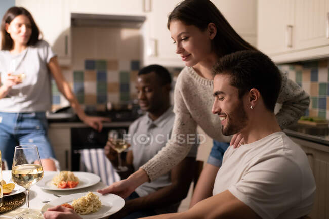 Jovem fêmea feliz colocando prato com prato saboroso perto namorado alegre durante a festa em casa com amigos diversos — Fotografia de Stock
