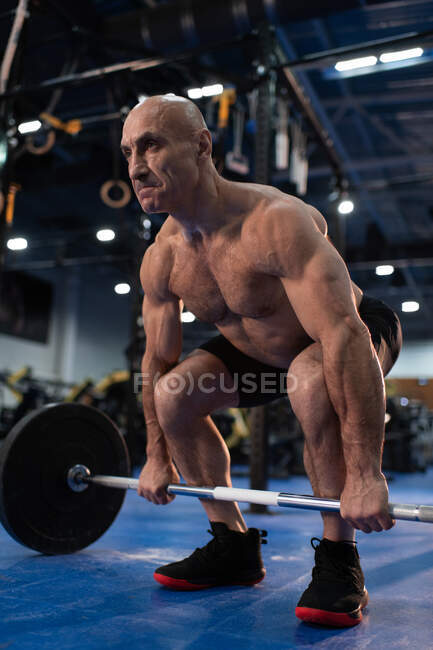 Full length concentrated elderly sportsman working out and deadlifting with heavy barbell in contemporary gym — Stock Photo