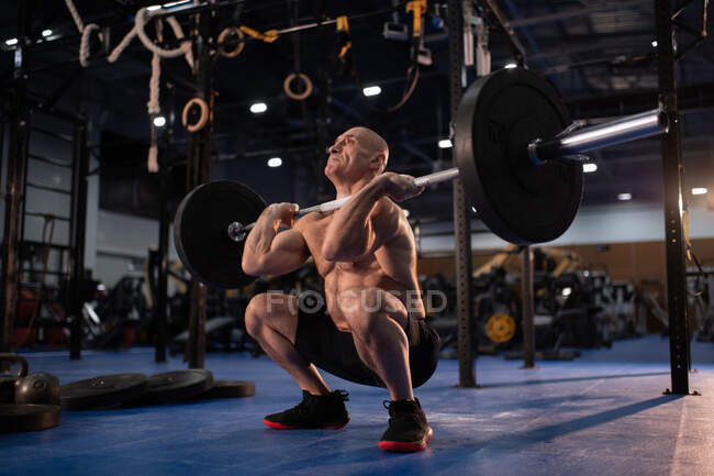 Full length bald elderly man in great shape doing front squat with heavy barbell while performing clean and jerk movement during weightlifting workout in contemporary gym — Stock Photo