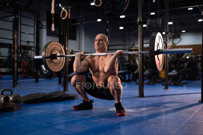 Longitud completa potente macho senior haciendo sentadilla frontal como parte de ejercicio limpio y tirón durante el entrenamiento intenso en el gimnasio - foto de stock