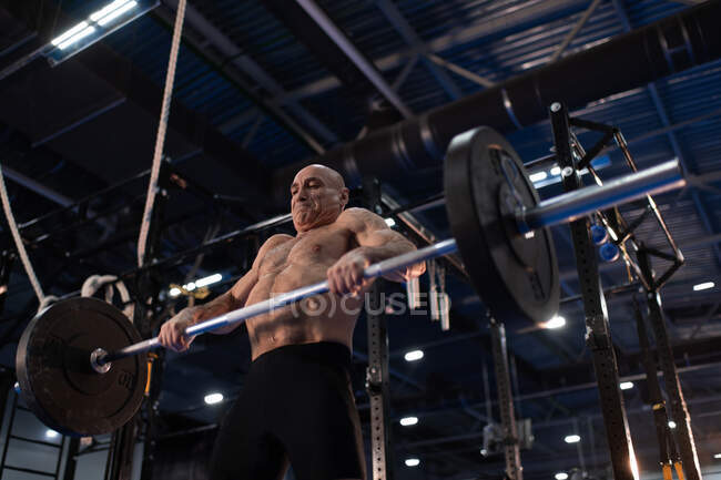 From below strong elderly man performing deadlift exercise with great effort while doing clean and jerk movement during intense weightlifting workout in modern gym — Stock Photo