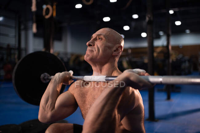 Bald shirtless elderly athlete squatting and doing clean and jerk exercise during weightlifting workout in contemporary gym — Stock Photo