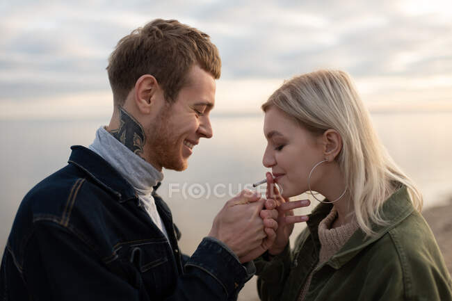 Side view cheerful man lighting marijuana joint for delighted woman while standing in evening countryside in autumn — Stock Photo