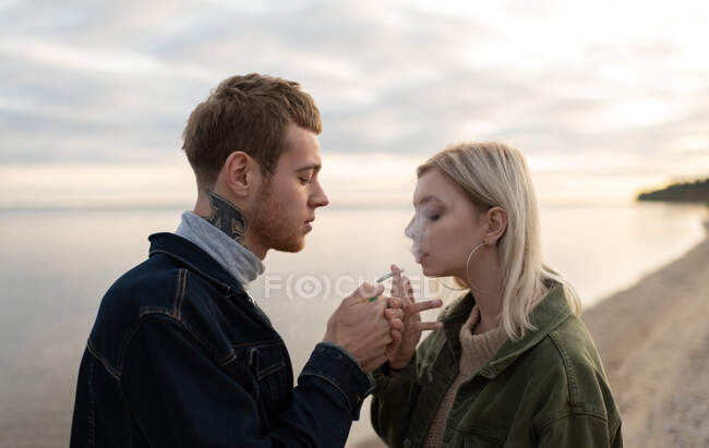 Vista lateral de jovens hipsters fumando maconha ao mesmo tempo em pé contra o lago calmo e céu nublado durante a data no campo de outono — Fotografia de Stock