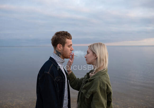 Side view of blonde woman giving cannabis cigarette to handsome tattooed boyfriend looking at each other on cold remote beach — Stock Photo