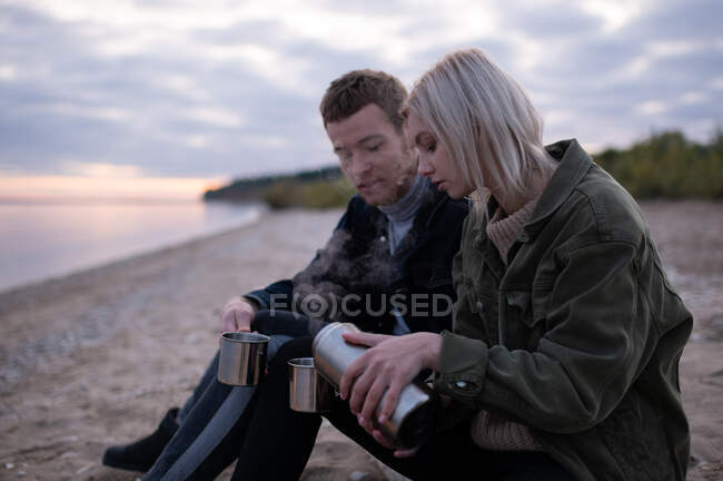 Side view of young blond woman pouring fresh aromatic tea into mugs while resting on lake shore with boyfriend during sundown — Stock Photo