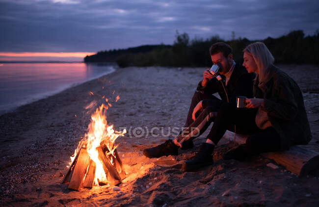 Vista lateral de los jóvenes hipsters que beben té caliente con malvaviscos mientras están sentados en el tronco cerca de la hoguera en llamas por la noche en el campo - foto de stock