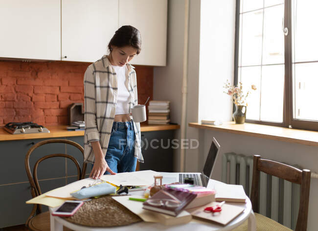 Mujer joven en casa tomando café y de pie en la mesa en la cocina leyendo notas mientras se prepara para el examen - foto de stock