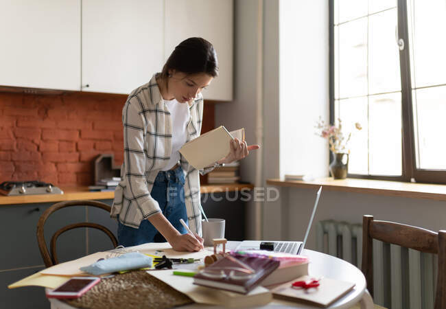 Зосереджена молода жінка в сорочці стоїть за столом на кухні і пише нотатки з підручника під час навчання вдома — стокове фото