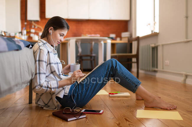Vista lateral de atractiva estudiante adolescente concentrada en auriculares sentados en el suelo preparándose para el examen en casa - foto de stock