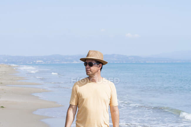 Hombre con sombrero y gafas de sol de pie en la orilla del mar, mientras mira hacia otro lado - foto de stock