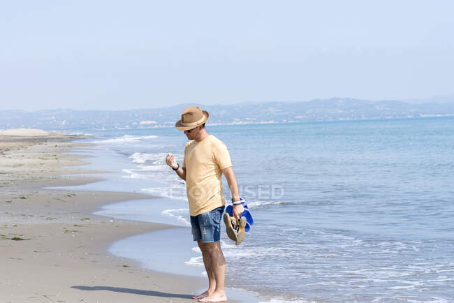 Man standing on seashore while holding shoes and observing a shell — Stock Photo