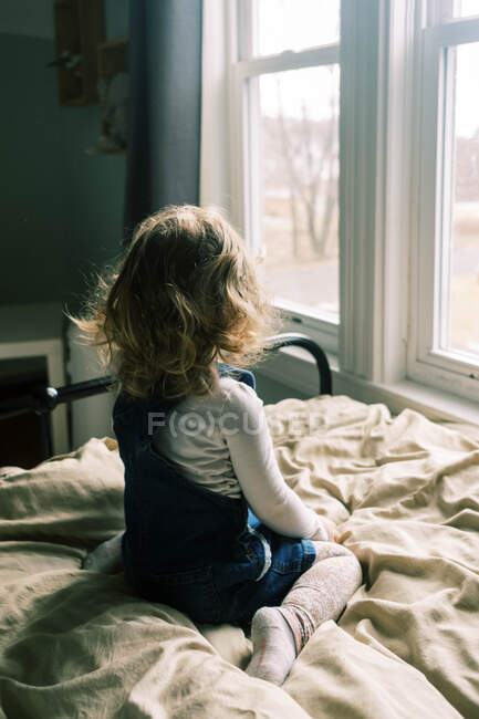 Piccola bambina seduta sul suo letto che guarda fuori dalla finestra — Foto stock