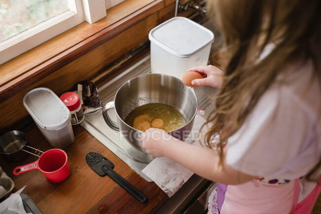 Menina vestindo um avental e quebrando ovos em uma tigela na cozinha — Fotografia de Stock