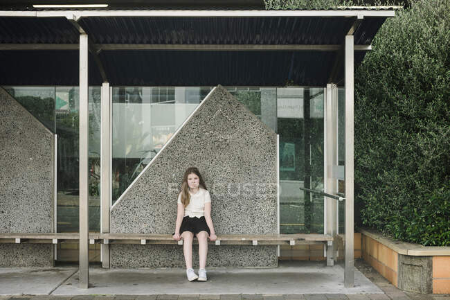 Young girl sitting alone on a wooden seat at a bus stop — Stock Photo