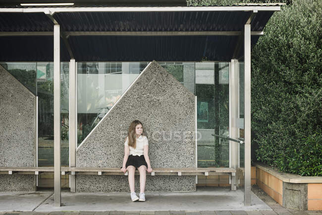 Молода дівчина сидить наодинці на дерев'яному сидінні на порожній зупинці автобуса — стокове фото